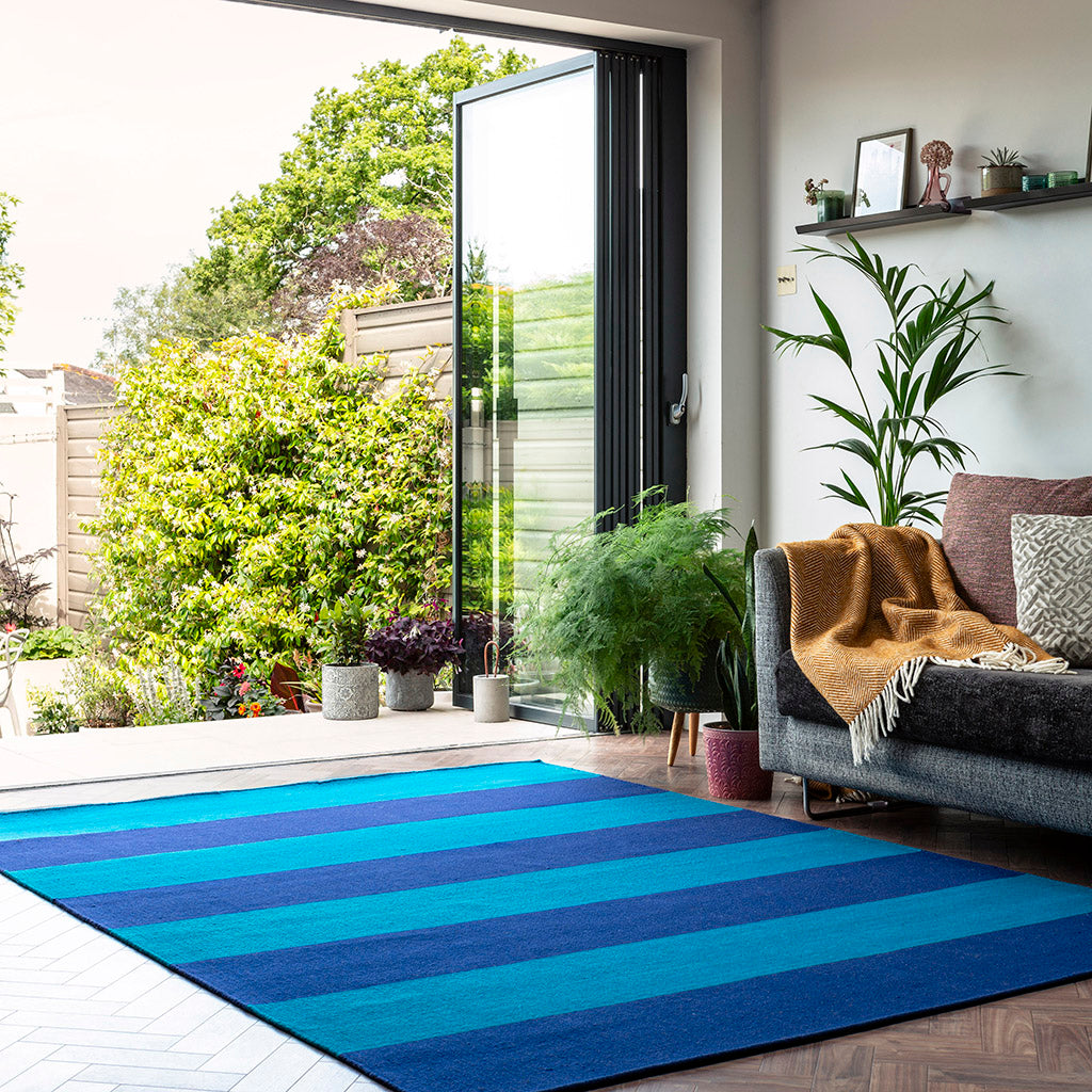 A sky blue and mid blue large striped wool rug in a living room with a sofa and bi-fold doors. This comes from a range of wooll rugs and runners which includes striped, flatweave and heavyweight styles made from ethically sourced New Zealand wool. Sizes include kitchen rugs, small bedroom rugs and long hallway runners